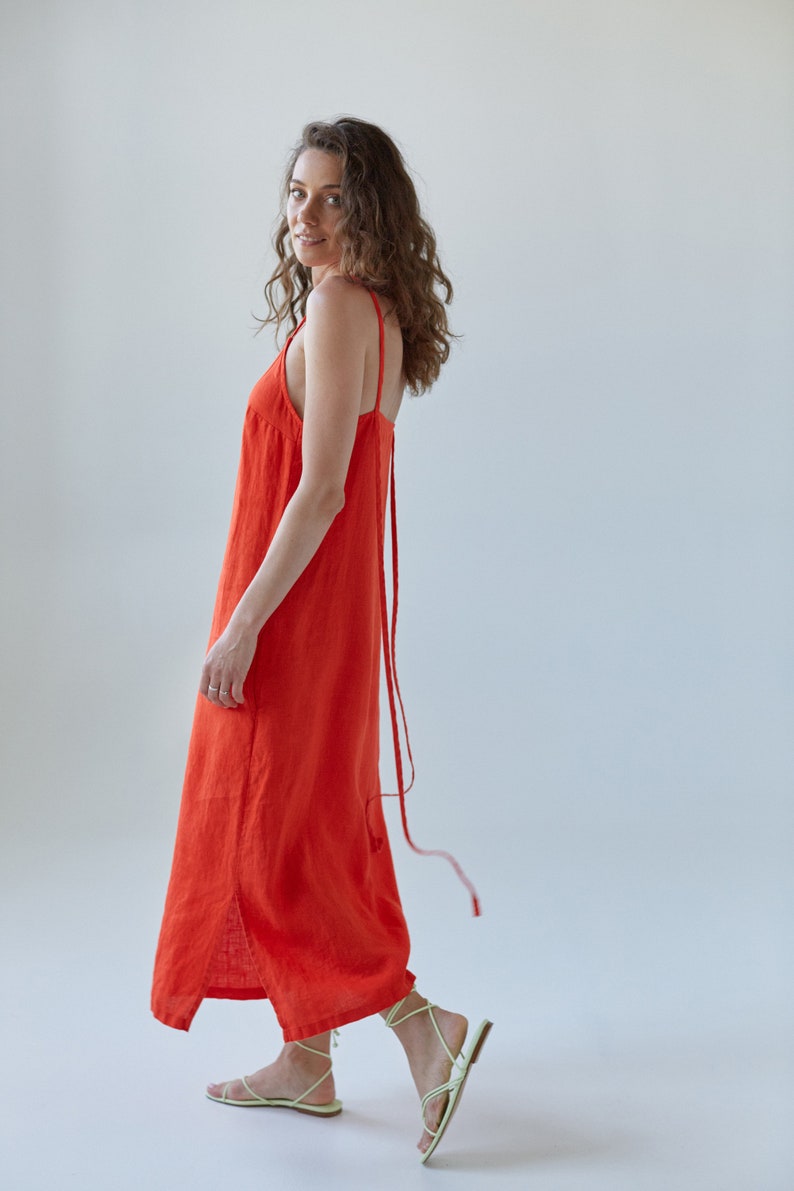May include: A woman wearing a bright orange maxi dress with thin straps. The dress has a side slit and a long tie that hangs down the back. She is wearing light green sandals.