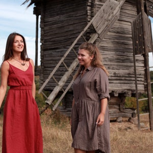 May include: Two women wearing long, flowing dresses stand in front of a wooden windmill. The woman on the left is wearing a red dress and the woman on the right is wearing a brown dress.