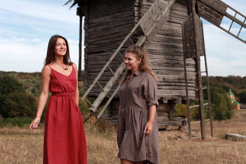 May include: Two women wearing long, flowing dresses stand in front of a wooden windmill. The woman on the left is wearing a red dress and the woman on the right is wearing a brown dress.