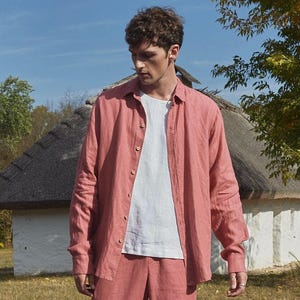 May include: A man wearing a coral linen shirt with a white linen undershirt and matching coral linen pants. He is standing in front of a white building with a thatched roof.