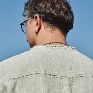 May include: A person with short, brown hair wearing a light beige linen shirt. The person is wearing a silver chain necklace and a silver hoop earring in their left ear.