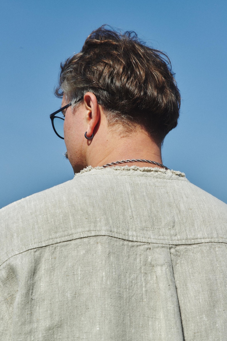 May include: A person with short, brown hair wearing a light beige linen shirt. The person is wearing a silver chain necklace and a silver hoop earring in their left ear.