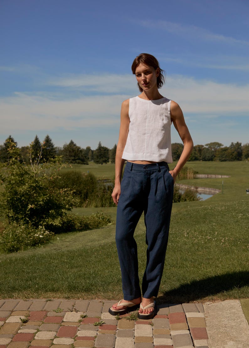 May include: A woman wearing a white sleeveless top and navy blue pants. The pants have a pleated front. She is wearing flip-flops and standing on a brick walkway. The background is a green field and blue sky.