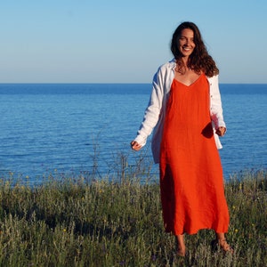 May include: A woman in a white linen shirt and a long orange dress stands in a field of green grass with a blue body of water in the background.