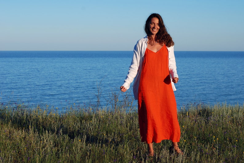 May include: A woman in a white linen shirt and a long orange dress stands in a field of green grass with a blue body of water in the background.