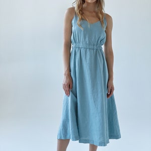 May include: A light blue linen dress with thin straps and a gathered waist. The dress is midi length and has a relaxed fit. The model is wearing white espadrille sandals.