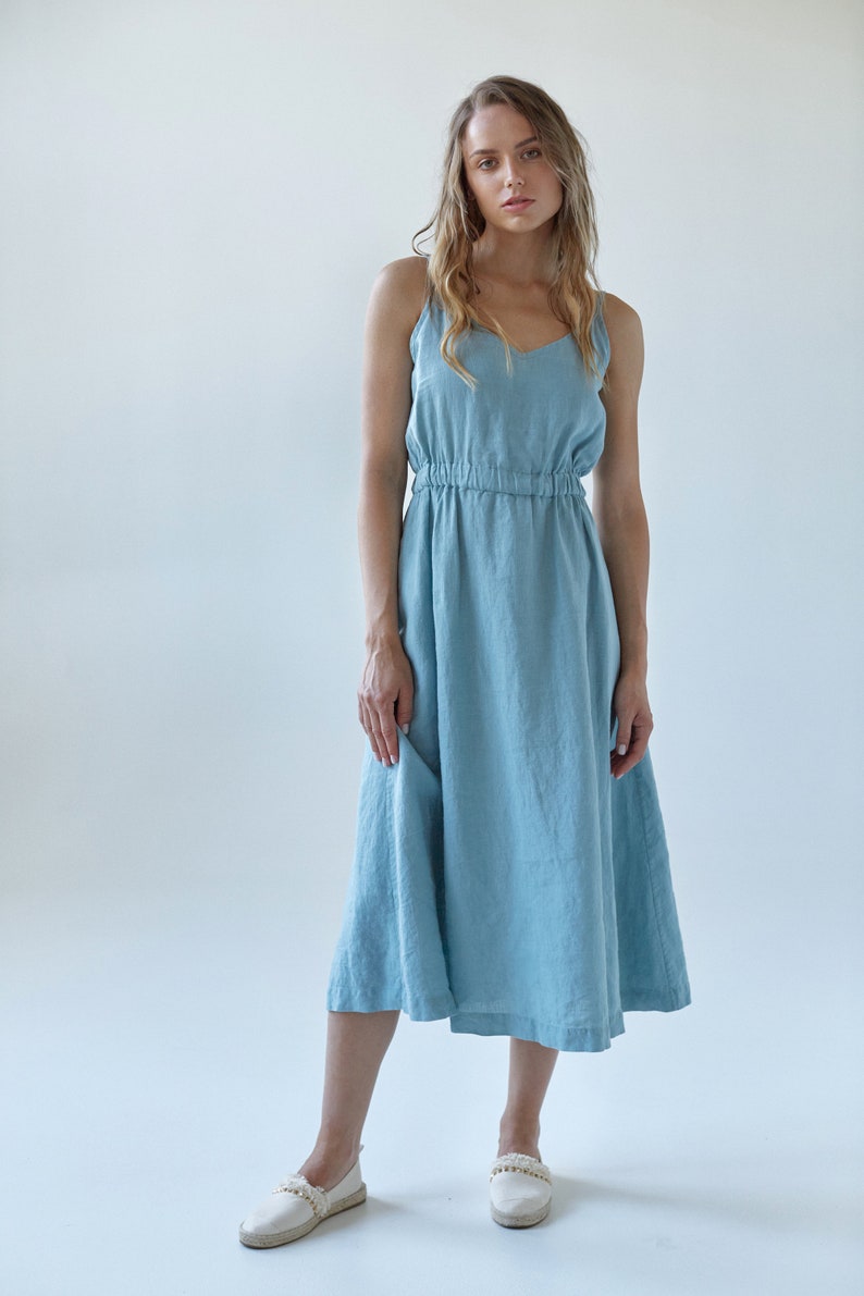 May include: A light blue linen dress with thin straps and a gathered waist. The dress is midi length and has a relaxed fit. The model is wearing white espadrille sandals.