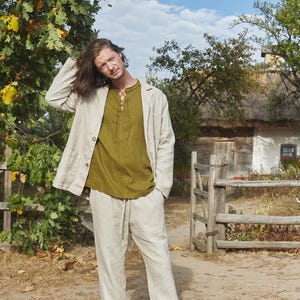 May include: A man wearing a light beige linen jacket over a green linen shirt and matching linen pants. He is standing in front of a wooden fence and a white building with a thatched roof.