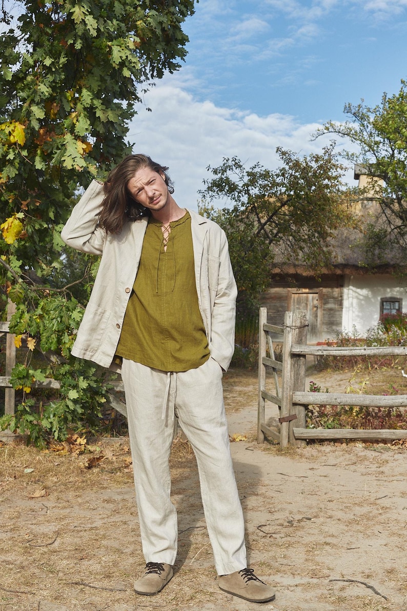 May include: A man wearing a light beige linen jacket over a green linen shirt and matching linen pants. He is standing in front of a wooden fence and a white building with a thatched roof.