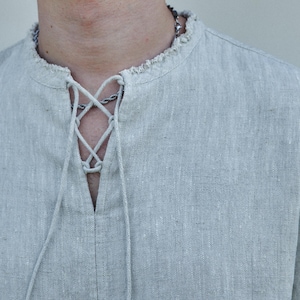 May include: A light gray linen shirt with a lace-up neckline. The shirt has a relaxed fit and a slightly frayed collar.