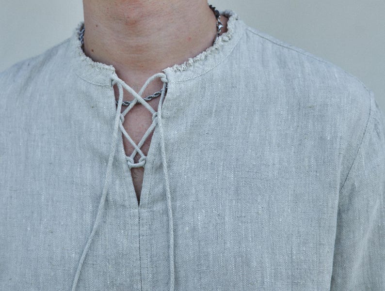 May include: A light gray linen shirt with a lace-up neckline. The shirt has a relaxed fit and a slightly frayed collar.