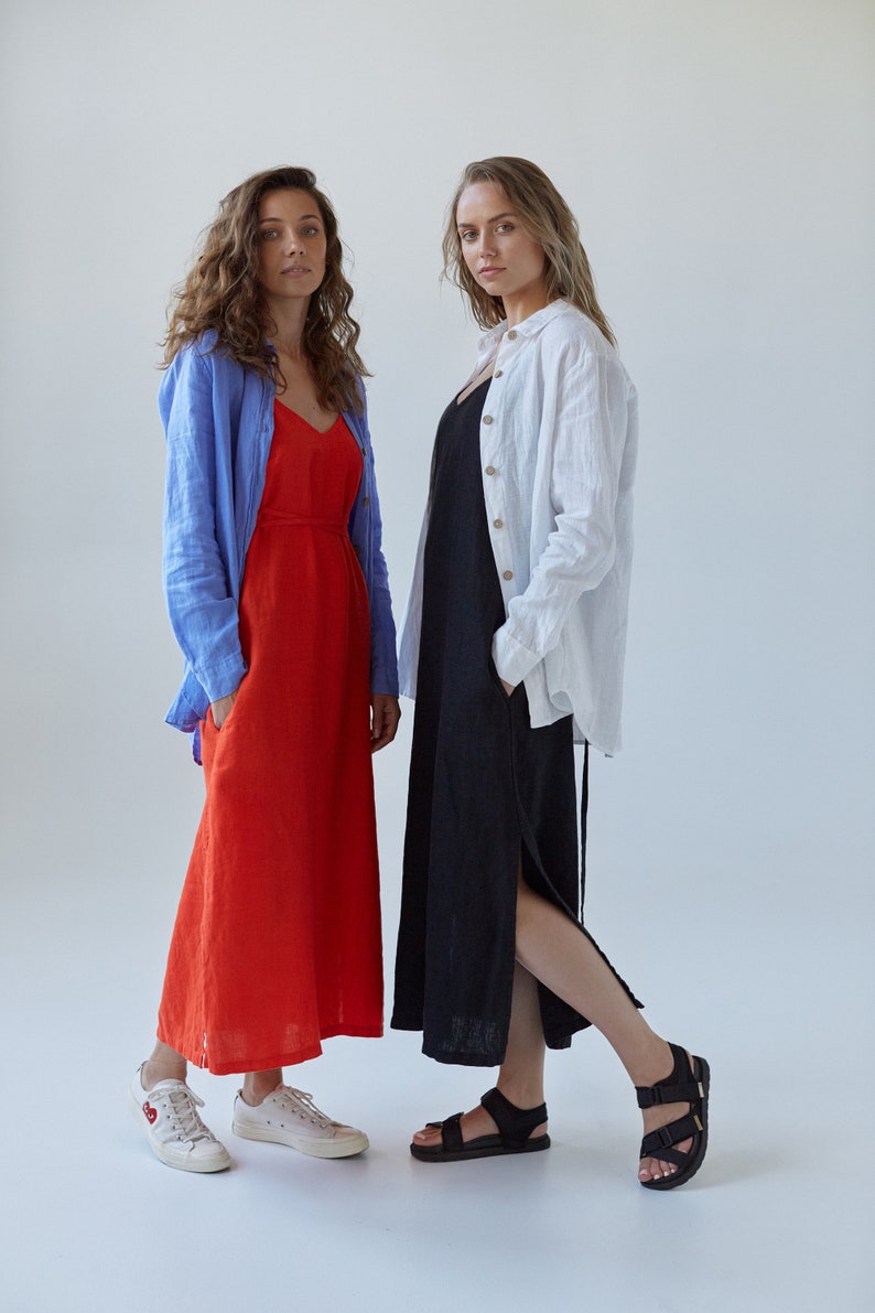 May include: Two women wearing long dresses. The woman on the left is wearing a red dress with a blue shirt over it. The woman on the right is wearing a black dress with a white shirt over it. Both women are wearing sandals.
