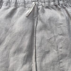 May include: Close-up of light gray linen shorts. The shorts have an elastic waistband with a white drawstring. The fabric has a textured appearance. The shorts are made of a lightweight material.