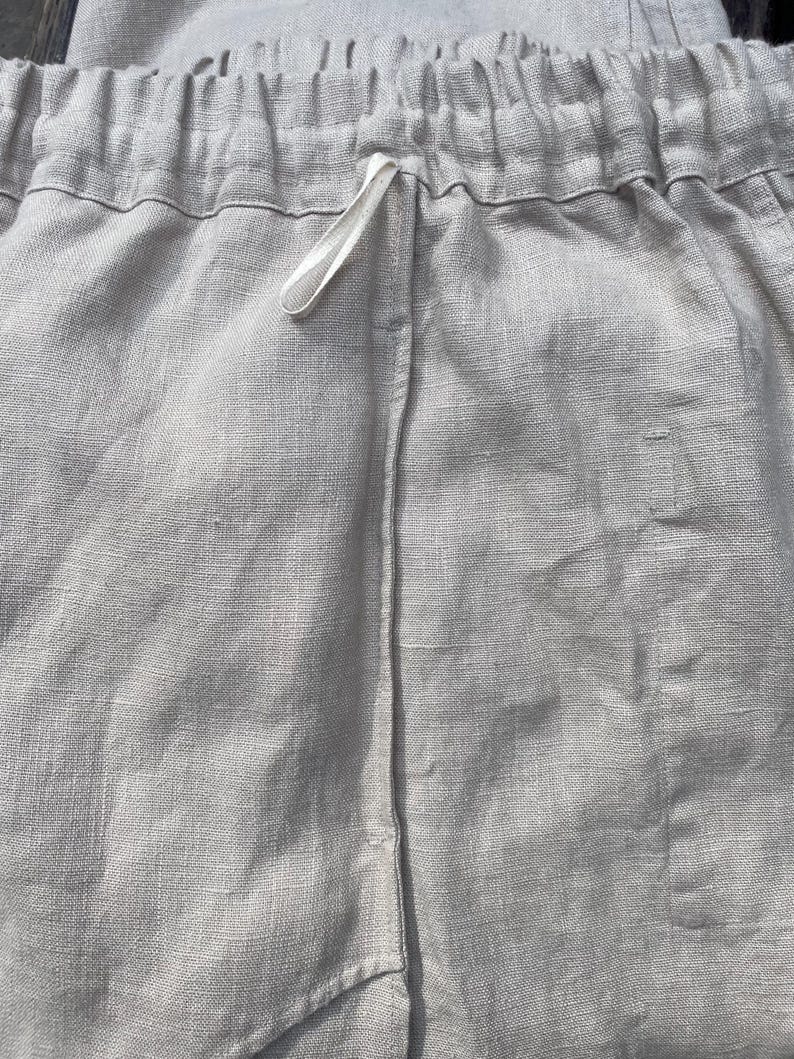 May include: Close-up of light gray linen shorts. The shorts have an elastic waistband with a white drawstring. The fabric has a textured appearance. The shorts are made of a lightweight material.