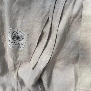 May include: Close-up of a light gray linen garment with visible seams and a small label. The label features a circular illustration and text, likely indicating the brand or origin. The fabric has a textured appearance.