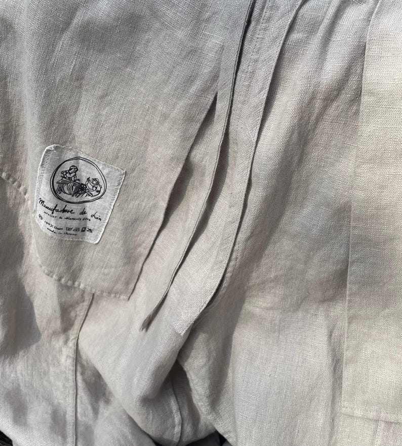 May include: Close-up of a light gray linen garment with visible seams and a small label. The label features a circular illustration and text, likely indicating the brand or origin. The fabric has a textured appearance.