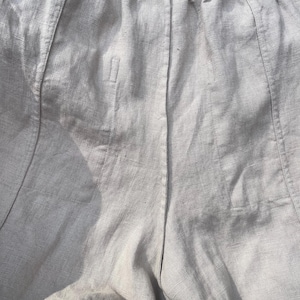 May include: Close-up of light gray linen pants. The pants have a drawstring waistband and vertical seams down the front and back. The fabric has a textured appearance. The pants appear to be lightweight and suitable for warm weather.