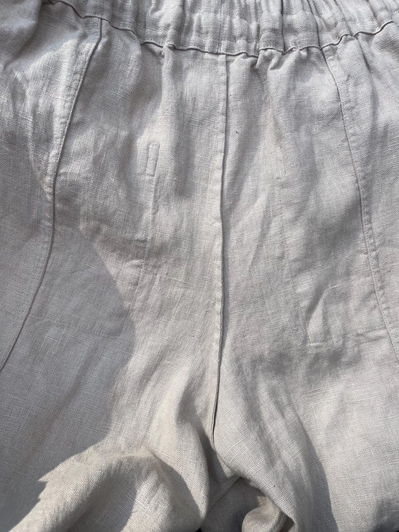 May include: Close-up of light gray linen pants. The pants have a drawstring waistband and vertical seams down the front and back. The fabric has a textured appearance. The pants appear to be lightweight and suitable for warm weather.
