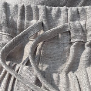 May include: Close-up of light beige linen pants. The image highlights the elastic waistband with a drawstring, showcasing the fabric's texture and the garment's construction. The focus is on the details of the pants.