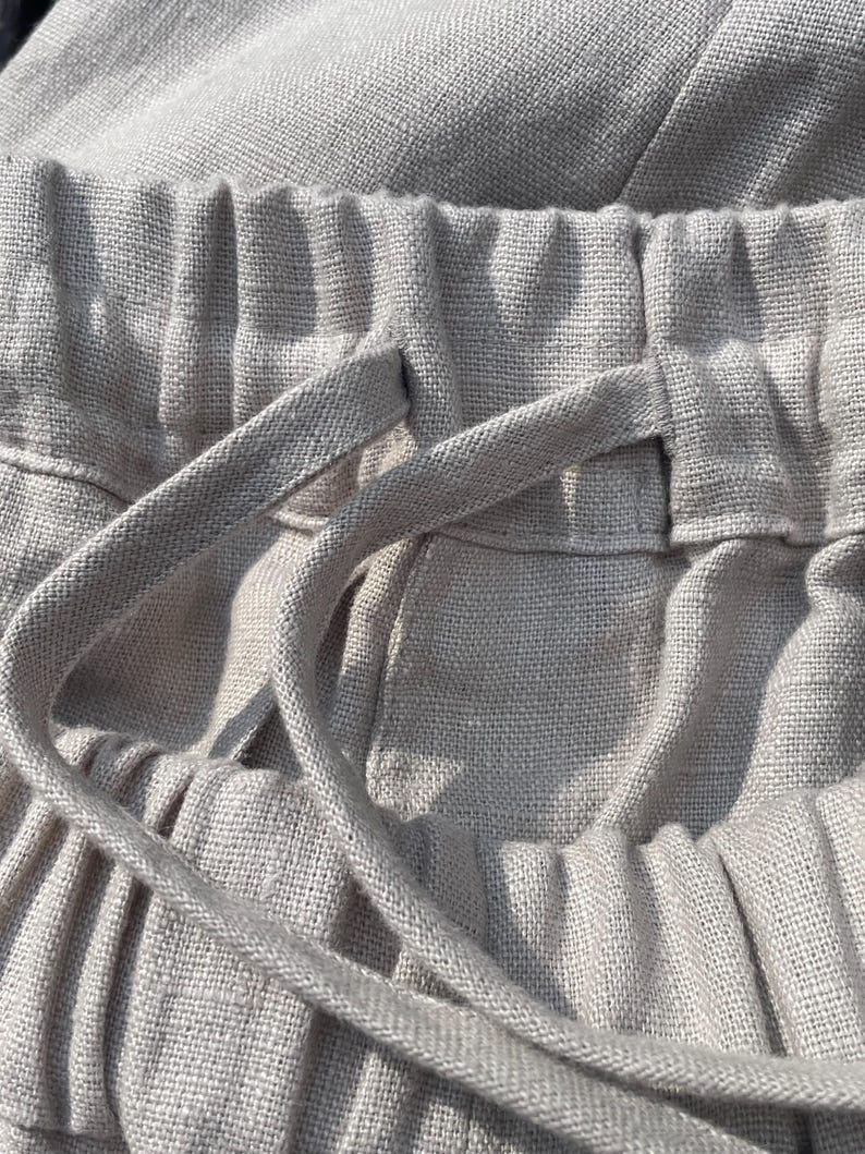 May include: Close-up of light beige linen pants. The image highlights the elastic waistband with a drawstring, showcasing the fabric's texture and the garment's construction. The focus is on the details of the pants.