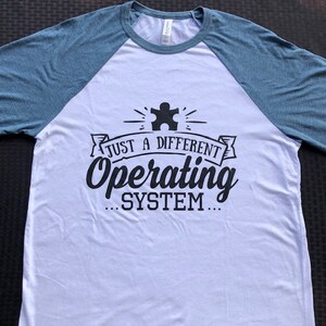 May include: A white and blue baseball tee shirt with a black graphic that says "Just a different operating system..." with a puzzle piece symbol above the text.