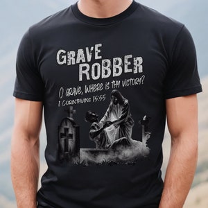 May include: Black t-shirt with a graphic of Jesus Christ rising from a grave. The text "Grave Robber" is at the top of the graphic, and the text "O grave, where is thy victory? 1 Corinthians 15:55" is below the graphic.