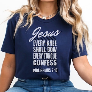 May include: Navy blue t-shirt with white text that reads "Jesus Every Knee Shall Bow Every Tongue Confess Philippians 2:10". The shirt is a short-sleeved crew neck.