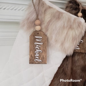 May include: White and brown faux fur Christmas stocking with a wooden tag that says "Michael" in white lettering. The tag is attached with twine and wooden beads.