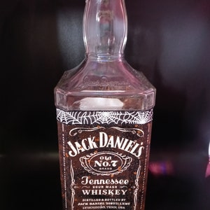 May include: A clear glass bottle of Jack Daniel's Tennessee Sour Mash Whiskey. The label is black with white text and a spiderweb design. The bottle is illuminated with colorful lights.