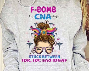 May include: Gray hooded sweatshirt with a graphic design of a woman with a messy bun wearing glasses and a tie-dye headband. The text reads "F-Bomb CNA" and "Stuck Between IDK, IDC and IDGAF".