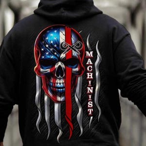 May include: Black hoodie featuring a skull design with an American flag motif. The skull is red, white, and blue, with the word "MACHINIST" printed vertically. The design includes a red stripe and silver flame-like accents.