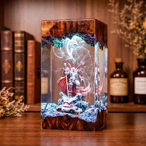 May include: A rectangular lamp featuring a dark wood base and top with a clear resin center. Inside the resin, a miniature figure with a red cape stands on a creature, surrounded by swirling white smoke effects and purple and blue accents.