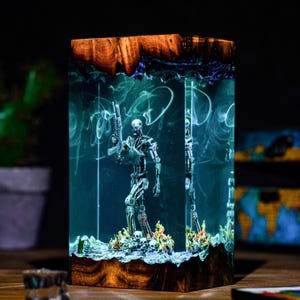 May include: A rectangular resin block featuring a metallic silver robotic skeleton figure holding a rifle, standing on a base of skulls and flames. The clear resin is illuminated with a glowing teal light and swirling smoke effects, framed by dark wood.