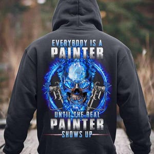 Könnte beinhalten: Schwarzer Kapuzenpullover mit Totenkopf-Design. Das Design zeigt einen blauen Totenkopf mit Farbspritzgeräten und den Text "EVERYBODY IS A PAINTER" und "UNTIL THE REAL PAINTER SHOWS UP".