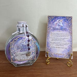 Op de afbeelding: Een glazen fles met het label "Unicorn Silk" met een paarse en blauwe vloeistof, een eenhoornafbeelding en tekst. Een decoratieve kaart met een eenhoornafbeelding en tekst staat op een gouden standaard. De fles is 50 ml.