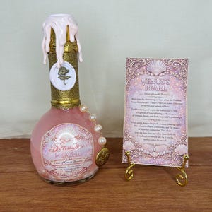 May include: A pink, round glass bottle with a gold glitter top and a white wax seal. The bottle has a label that reads "Venus's Pearl Elixir of Love & Beauty." A card with text and a gold stand is next to the bottle.