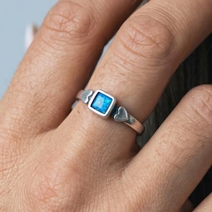 May include: A silver ring with a square, light blue gemstone at the centre. The ring features two small heart-shaped designs on either side of the gemstone. The ring is worn on a person's finger.