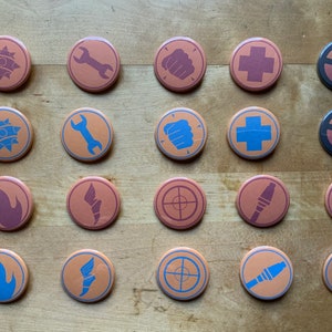 May include: A collection of twenty round buttons with various symbols in shades of blue and maroon on an orange background. The symbols include a wrench, fist, cross, and flame. The buttons are arranged on a wooden surface.
