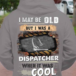 May include: Gray hoodie with the text "I MAY BE OLD BUT I WAS A DISPATCHER WHEN IT WAS COOL." The design features a headset graphic and a brick wall background.