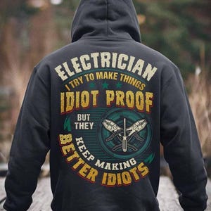Op de afbeelding: Donkergrijze hoodie met de tekst "ELECTRICIAN I try to make things idiot proof but they keep making better idiots". Het ontwerp bevat gereedschap en sterren in een cirkelvormig embleem. De hoodie heeft een capuchon met trekkoord.