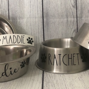 May include: Two stainless steel dog bowls with paw print decals. One bowl has the name "Maddie" on it, and the other bowl has the name "Ratchet" on it.