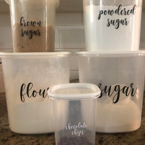 May include: Five clear plastic containers with labels for baking ingredients. The containers are labeled "brown sugar", "powdered sugar", "flour", "sugar", and "chocolate chips".