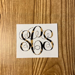 May include: A white sticker with a black monogram in a cursive font. The monogram is filled with a leopard print pattern in shades of brown and black.