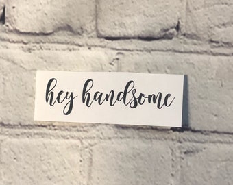 May include: A white rectangular card with the words "hey handsome" in elegant black cursive script. The card is set against a textured, light gray brick wall background.