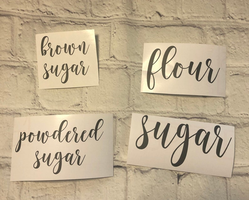 May include: Four white labels with black cursive text on a white brick background. The labels read "brown sugar", "flour", "powdered sugar", and "sugar".