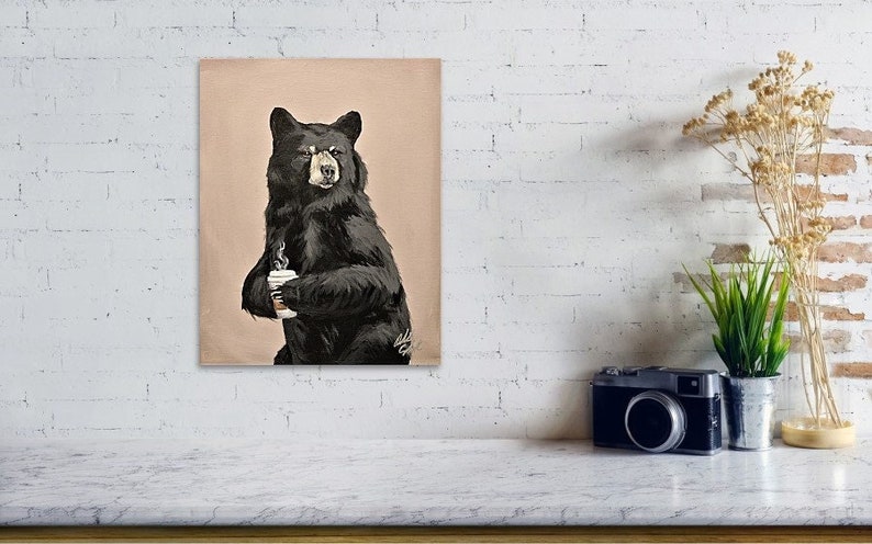 May include: A black bear sitting on a light brown background, holding a white cup with a brown drink. The bear is looking at the viewer.