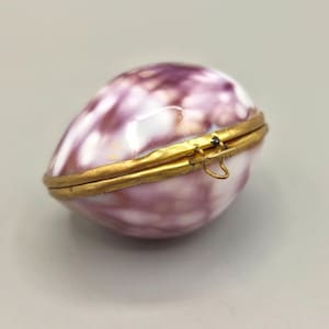 May include: A small, oval-shaped trinket box crafted from a polished, mottled purple and white seashell. The two halves of the shell are joined by a gold-toned metal hinge and secured with a simple wire clasp mechanism on the side.