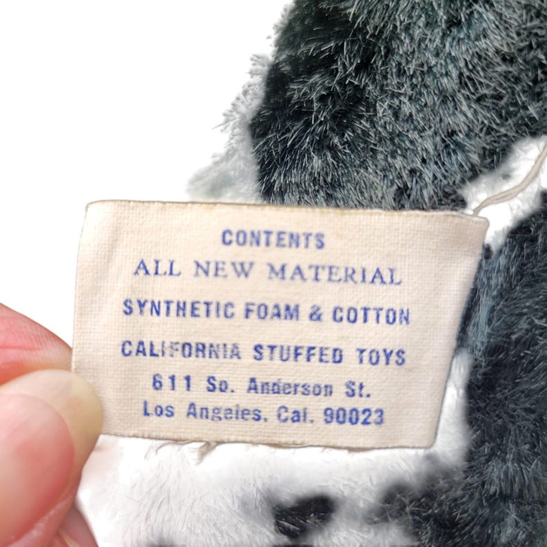 Puede incluir: Una etiqueta blanca con texto azul que dice "CONTENTS ALL NEW MATERIAL SYNTHETIC FOAM & COTTON CALIFORNIA STUFFED TOYS 611 So. Anderson St. Los Angeles. Cal. 90023"