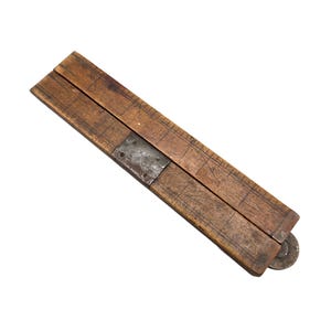 May include: A vintage wooden tool with a metal hinge and a small wheel at the end. The wood has a warm brown tone with visible grain and wear marks, suggesting age and use. The metal components show signs of oxidation.