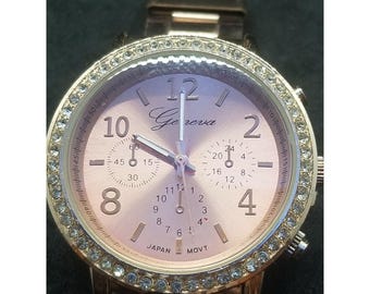May include: A rose gold Geneva watch with a crystal-studded bezel. The watch face is a light pink color with gold numerals and hands. The watch has three sub-dials and the text "Geneva" is printed on the watch face.